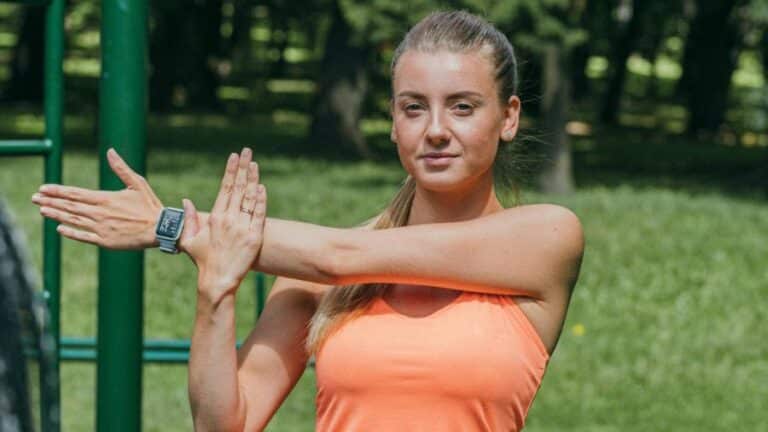 Mulher jovem usando smartwatch enquanto faz alongamento ao ar livre, em um parque com vegetação ao fundo