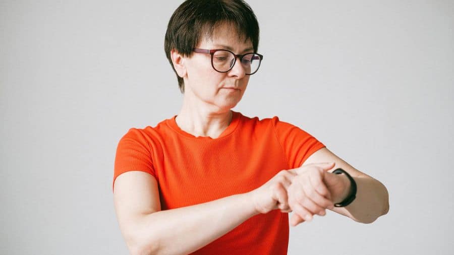 Mulher de óculos e camiseta laranja verificando as funções do smartwatch em um ambiente interno com fundo claro