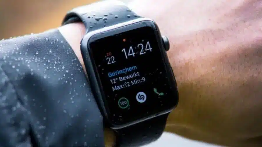 Smartwatch preto no pulso de uma pessoa com gotas de água na tela e na jaqueta, exibindo informações de clima e horário