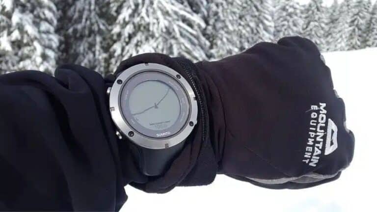 Smartwatch com GPS no pulso de uma pessoa usando luvas pretas em um ambiente com neve e árvores ao fundo