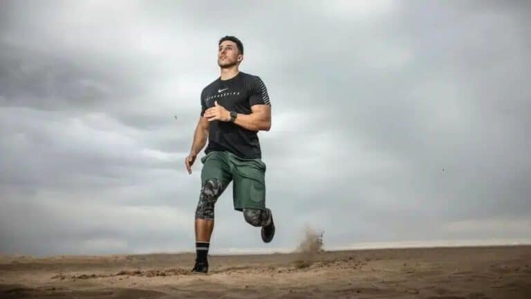 Homem correndo na areia usando roupa esportiva e relógio GPS, com céu nublado ao fundo