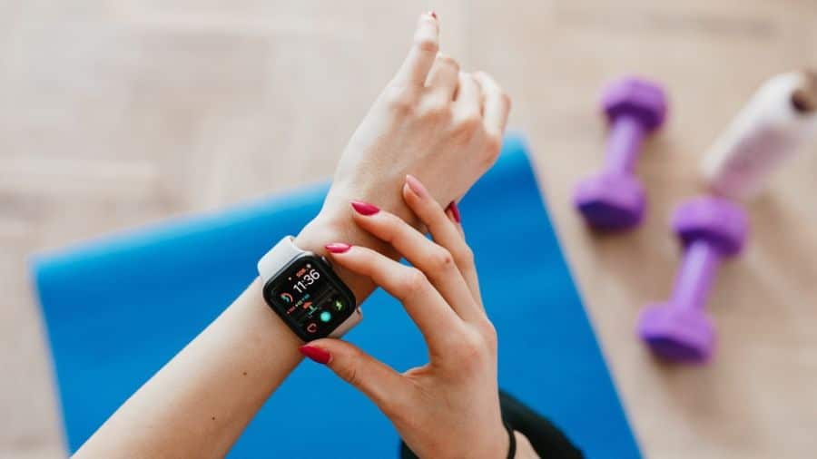 Pessoa ajustando smartwatch durante treino em ambiente interno, com colchonete e halteres ao fundo, usada como imagem ilustrativa do artigo