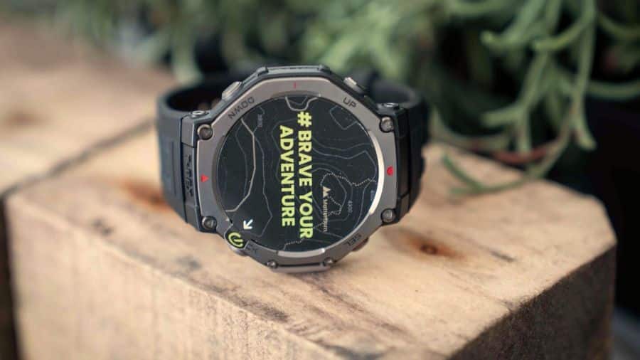 Amazfit T-Rex 3 exibindo a tela Brave Your Adventure sobre madeira com plantas ao fundo.